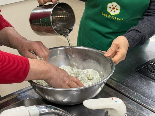調理室の作業台でエプロンを着た参加者たちがボウルに水を加えながら手で混ぜている写真