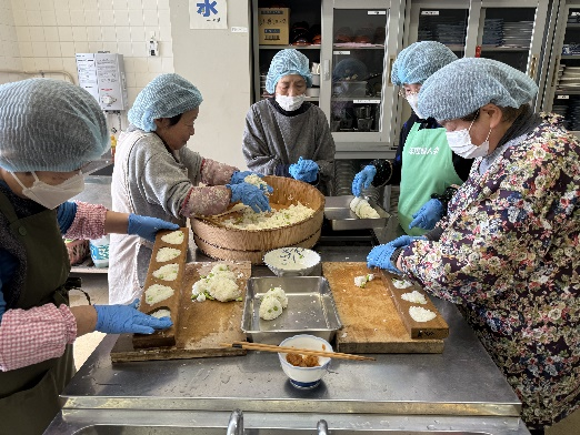 ヘアキャップと手袋を着用した女性たちが大きな木桶のご飯を分けておにぎりを作っている写真