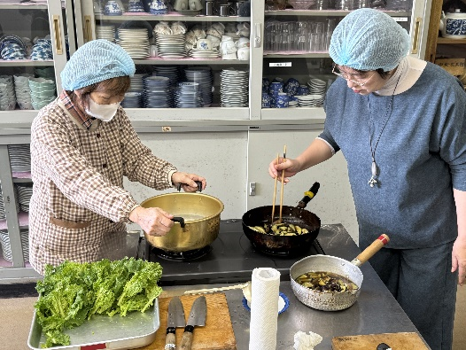 調理室でヘアキャップをかぶった女性たちが鍋とフライパンで調理しながら隣のバットの野菜を準備している写真