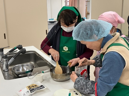 調理室の流し台の前でエプロンを着た女性たちが鍋の中の生地を木べらで混ぜながら材料を加えている写真
