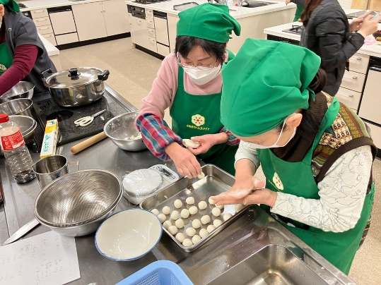 調理室の作業台で緑のバンダナをした女性たちが生地を手のひらで丸めてバットに並べている写真