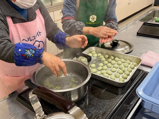 調理台のIHコンロの上でピンクのエプロンを着た参加者が丸めた団子を鍋に入れ、隣で別の参加者が団子を成形している写真