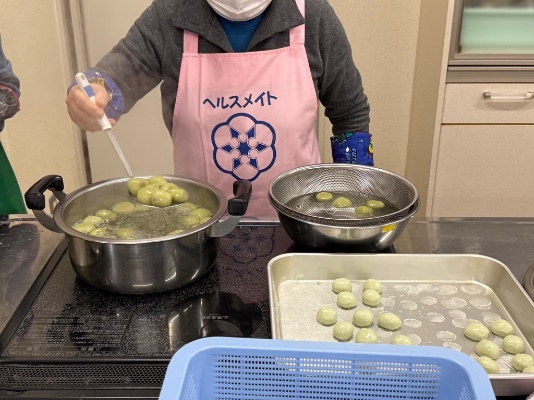 調理台の鍋の前でピンクのエプロンを着た参加者が茹で上がった団子をおたまで掬ってざるに移している写真