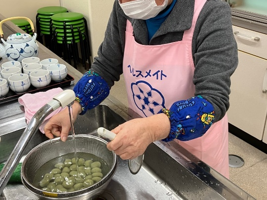 調理室の流し台でピンクのエプロンを着た参加者がざるに上げた団子に水をかけて冷ましている写真