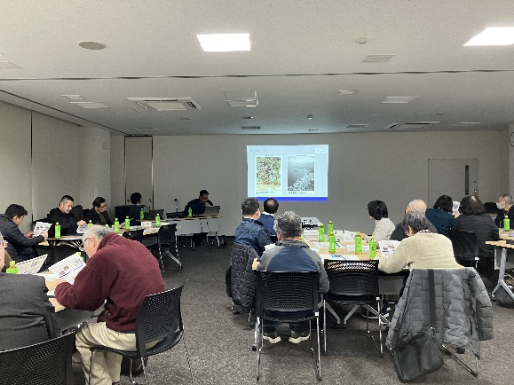 会議室でスクリーンのスライド説明を聞きながら資料を見る参加者たちの写真