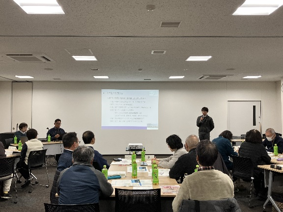 会議室前方で男性講師がマイクを持って発表している写真