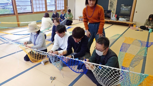 畳の部屋で参加者たちが横一列に座り、カラフルな網を編み進めているワークショップの写真