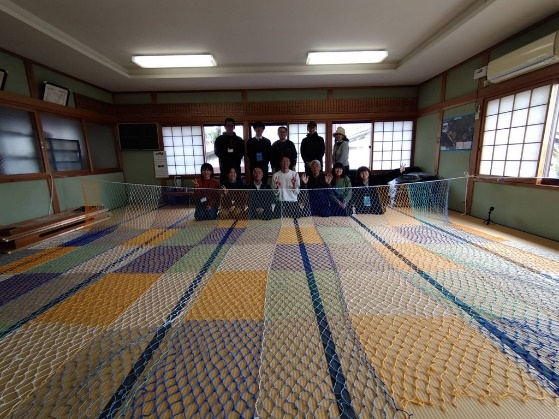 畳一面に完成した大きなカラフルな網を広げ、その奥で参加者全員が並んで記念撮影している写真