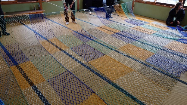 畳の上に広げた大きなカラフルな網の紐を、参加者たちが部屋の壁に繋げている写真