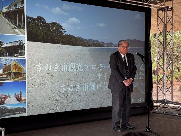 さぬき市観光プロモーションイベントで来賓が大型スクリーンの前に立って挨拶している写真