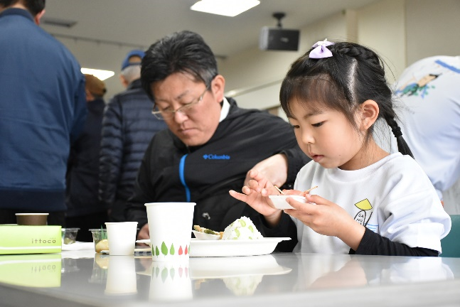 会場のテーブル席で男性と女の子が並んで料理を試食している写真