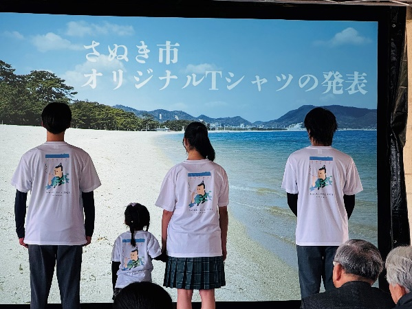 さぬき市オリジナルTシャツを着た子どもと若者がスクリーン前に背を向けて並んでいる写真