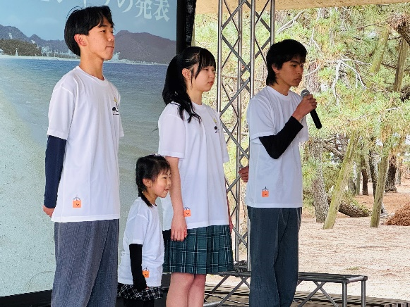 さぬき市オリジナルTシャツを着た子どもと若者がステージ上に並び、一人がマイクで話している写真