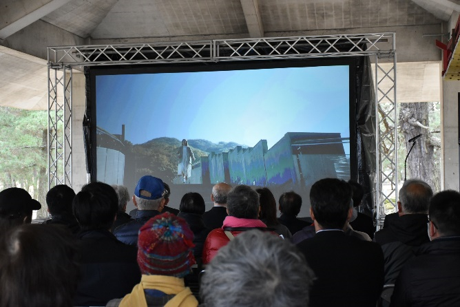 屋根付きの会場に設置された大型スクリーンの映像を大勢の観客が見ている写真