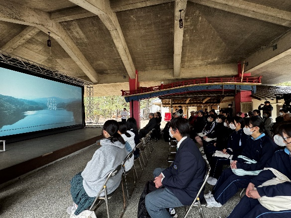 屋根付きの会場で椅子に座った参加者たちがスクリーンに映された映像を見ている写真