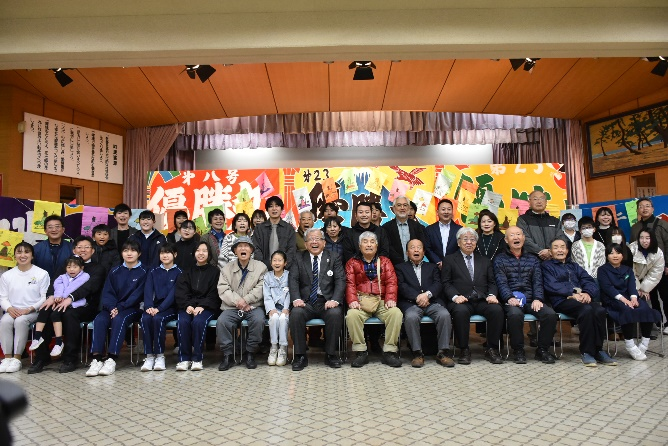 カラフルな背景幕の前で子どもから大人まで多世代が横一列に並び記念撮影している写真