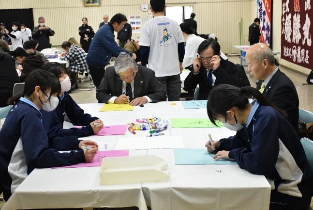 会場内のテーブルで中学生と地域の大人が布にメッセージを書いているワークショップの写真