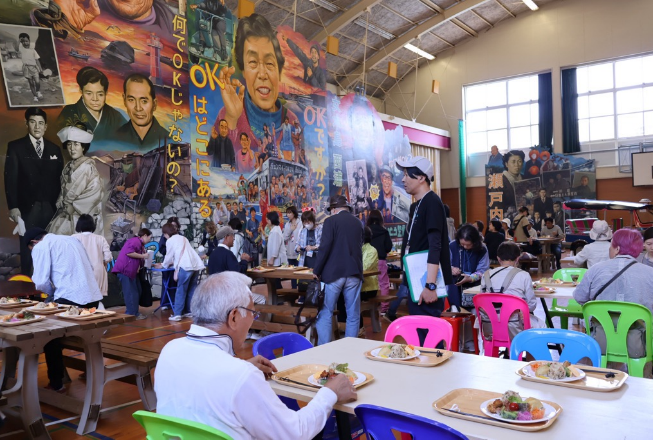 壁一面に大きなポスターが沢山貼られた会場で食事を楽しむ参加者の写真