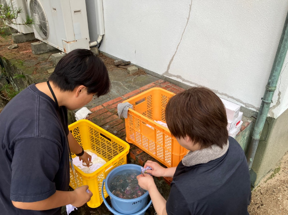 屋外でバケツの中のレンズを洗って拭いている二人の写真
