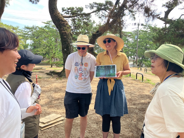 松林の中で帽子をかぶったアーティストがタブレットの画面を見せながら参加者と話している写真