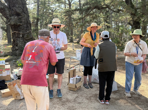 松林の中で帽子をかぶったアーティストと参加者たちがテーブルの前で交流している写真
