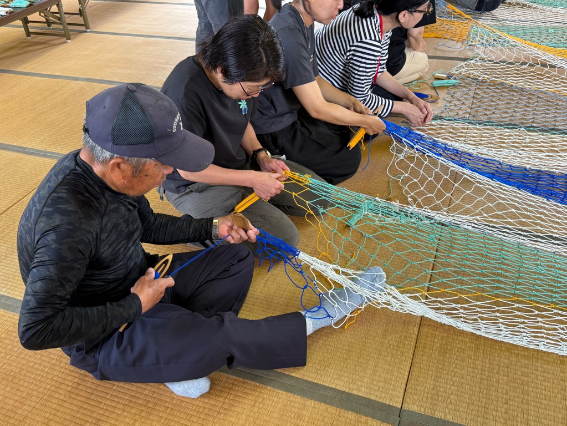 畳の部屋で帽子をかぶった男性と参加者たちがカラフルなネットを並んで編んでいる写真