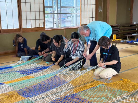 畳の部屋で指導役の男性が参加者たちと一緒にカラフルなネットを編んでいる写真