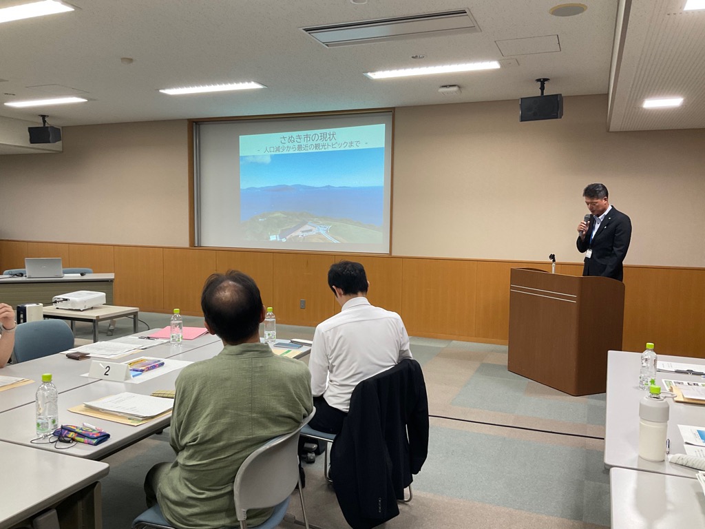 会議室にて挨拶を行っている男性と挨拶を聞く参加者の写真
