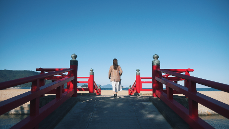 赤い橋の上を海に向かって進む女性の後ろ姿を捉えた写真
