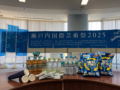 瀬戸内国際芸術祭2025の青い案内パネルの前に飲み物とスナック菓子が並べられている写真
