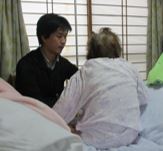 ベッドの上で座る高齢女性を支える男性の写真