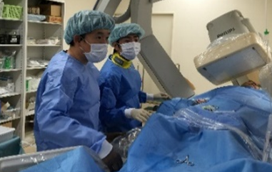 手術着姿の2人が青い布をかけて横になった人のわきに立っている写真