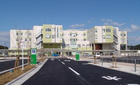 道路と駐車場の奥に4階建の白い壁をした建物が建っている写真