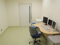 白い壁に囲まれた部屋の壁際に3枚のモニターとキーボードが置かれているデスクが設置されている写真