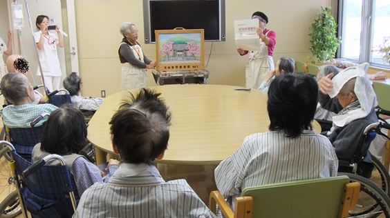 丸テーブルを囲んで座る高齢の男女に向かって紙芝居を行う女性の写真