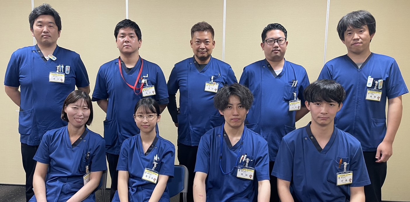 青いスクラブを着た男女9人が2列に並んだ集合写真