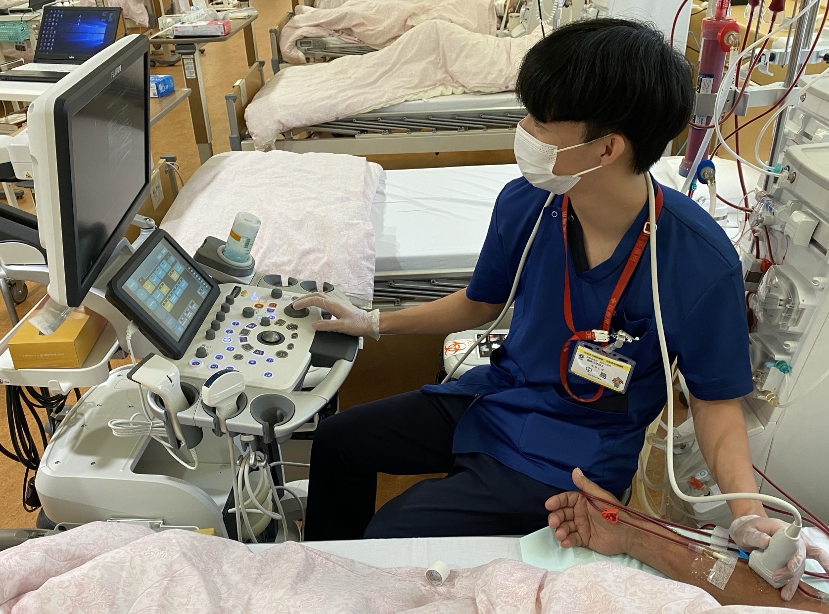 横たわる人の腕に機材を当てながらモニターを見る青いスクラブ姿の男性の写真