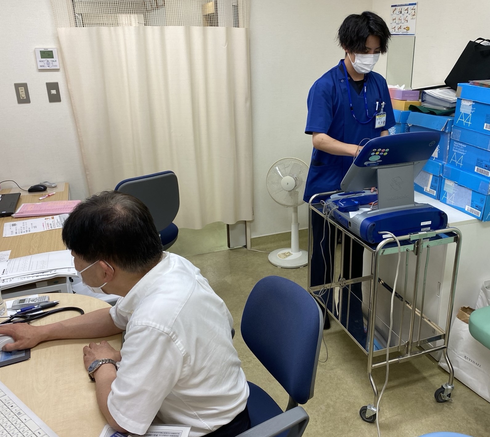 座っているワイシャツ姿の男性の斜め後ろで青いスクラブ姿の男性がモニターを操作している写真