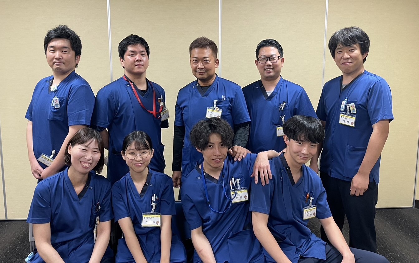青色のスクラブを着た男女9人の笑顔の集合写真
