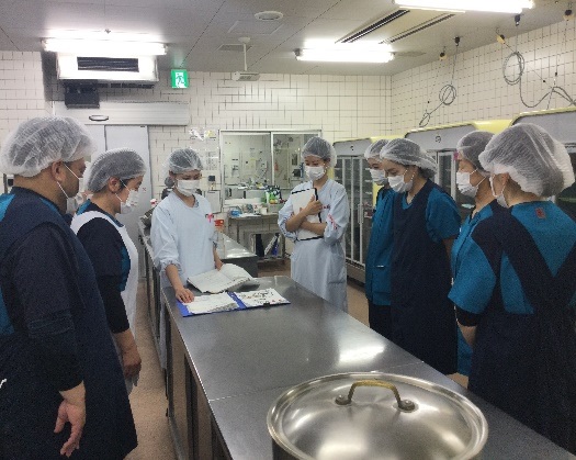 調理机の周りで衛生キャップやエプロンを付けた人たちが説明を聞いている写真