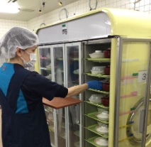 衛生キャップやエプロンを付けた人がガラスのついた棚に盛り付けをした皿を入れている写真