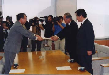 カメラがたくさんある前で書状を受け渡ししている男性の写真