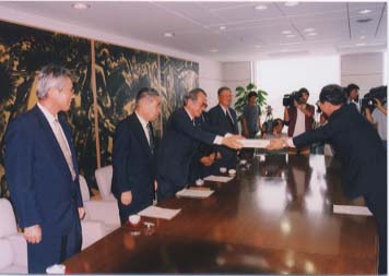 机の両側に立ち書状を受け渡している男性たちの写真