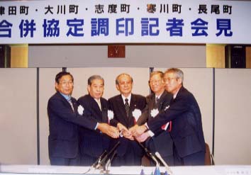 合併協定調印記者会見と書かれた横断幕の前で手を取り合っている5名の男性の写真