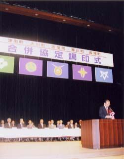 合併協定調印式と横断幕が掲げられた下で話す人の写真