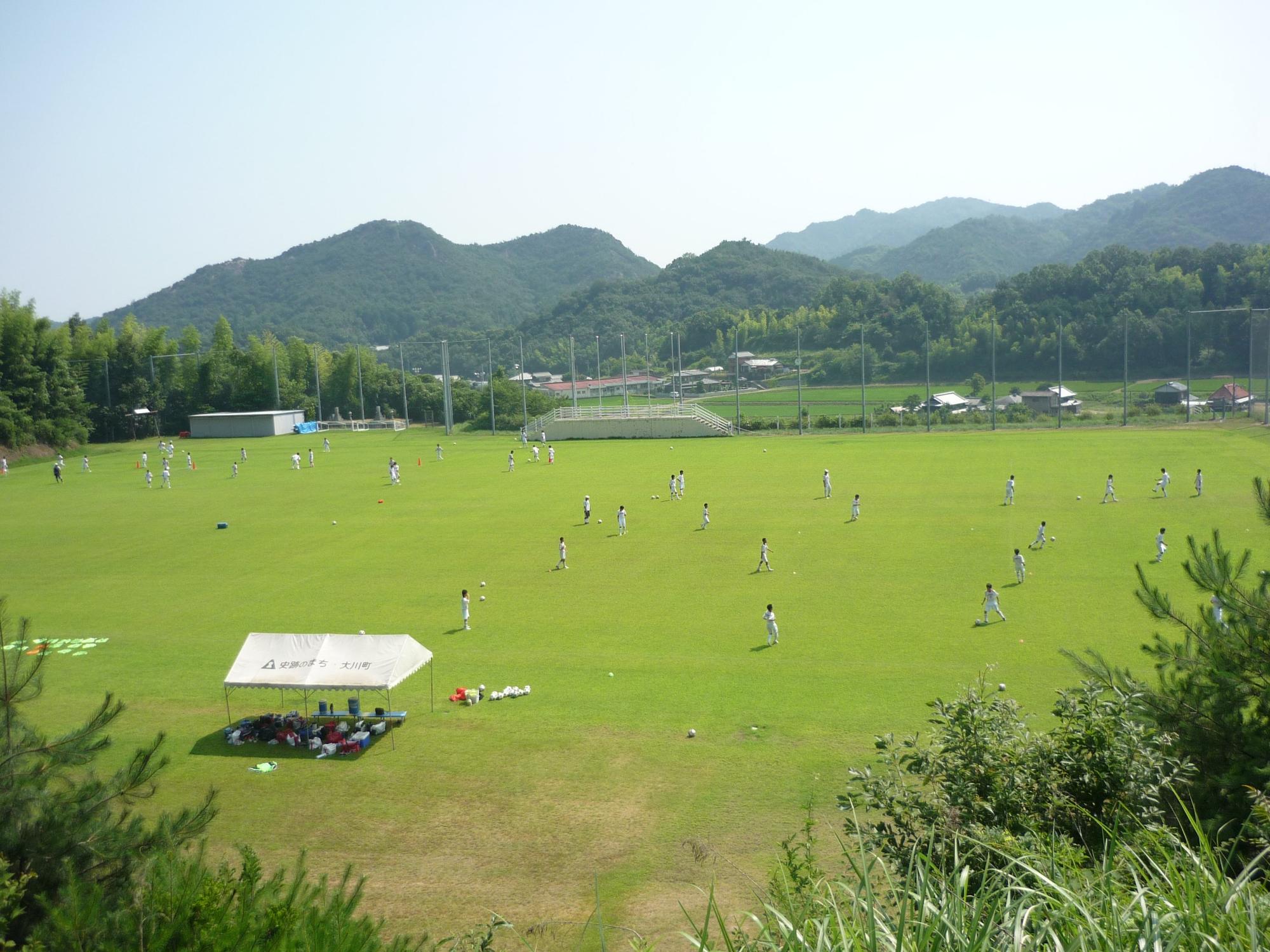 周りを山に囲われた広い芝生のグラウンドでサッカーをしている写真