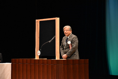 グレーのスーツを着用したメガネ姿の男性が演台に立ち、話している写真