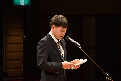 スーツを着用した男性が折りたたまれた紙を手にマイクの前で話している写真