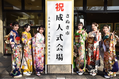 「祝 さぬき市成人式会場」と書かれた看板の左右に立つ振り袖姿の女性たちの写真