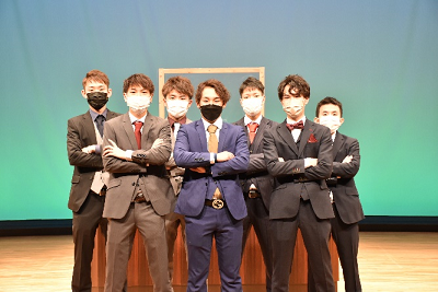 スーツ姿の7人の男性が2列に並び腕を組んでいる写真